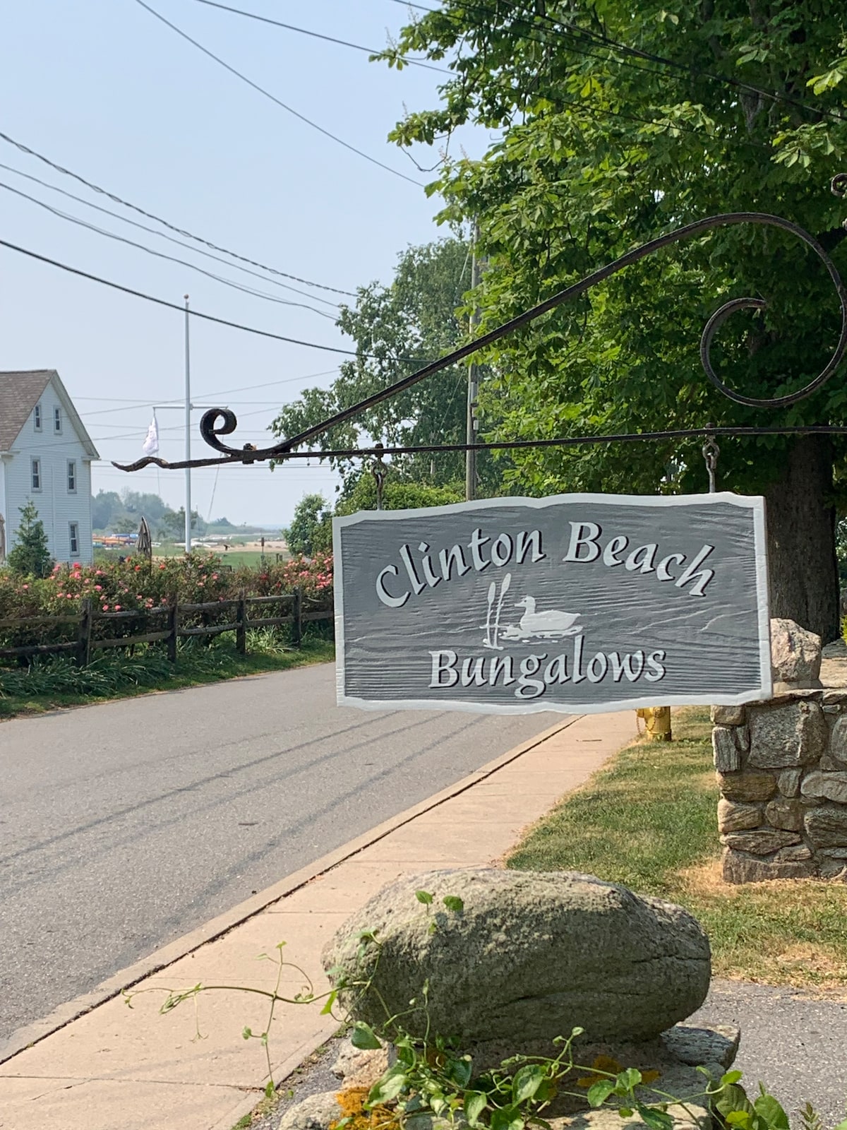 A welcoming wooden sign marks the entrance to Clinton Beach Bungalows, suspended from an ornate bracket. The sign features elegant lettering and an image of a bird, surrounded by lush greenery and a view of the nearby street lined with trees.