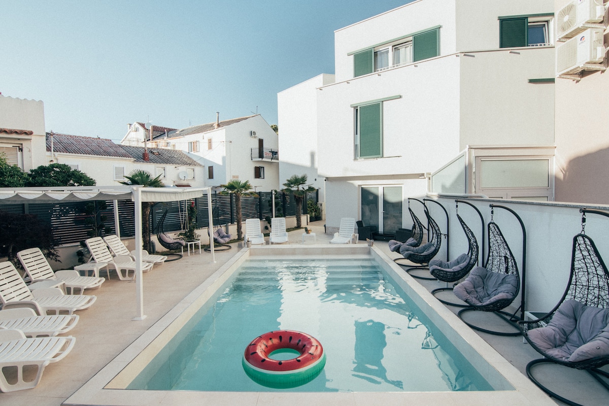 A serene pool area features a rectangular swimming pool surrounded by several lounge chairs and unique hanging chairs. A floating inflatable ring rests in the water, while shaded areas are provided for relaxation. The backdrop includes white buildings and greenery.