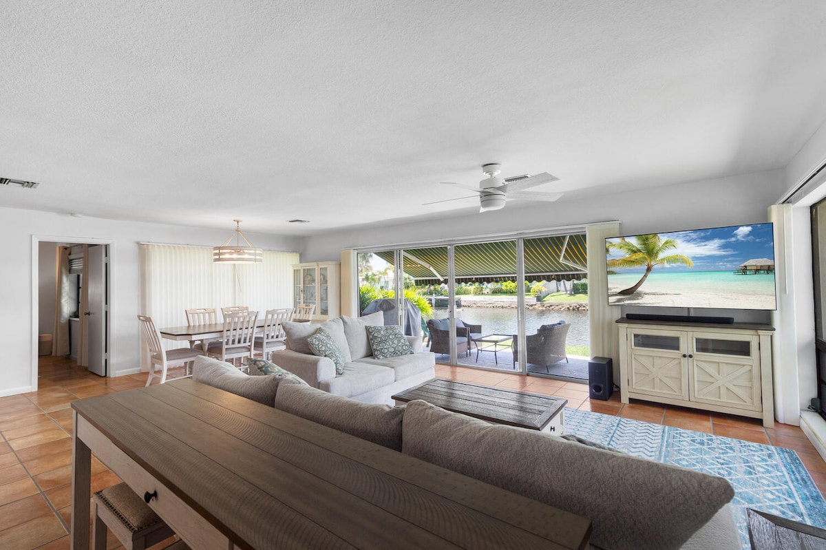 The open-concept living area features a spacious couch and dining table, both offering views of the serene waterfront through large sliding glass doors. A neutral color palette and natural light create a calm environment, while a ceiling fan adds comfort to the space.