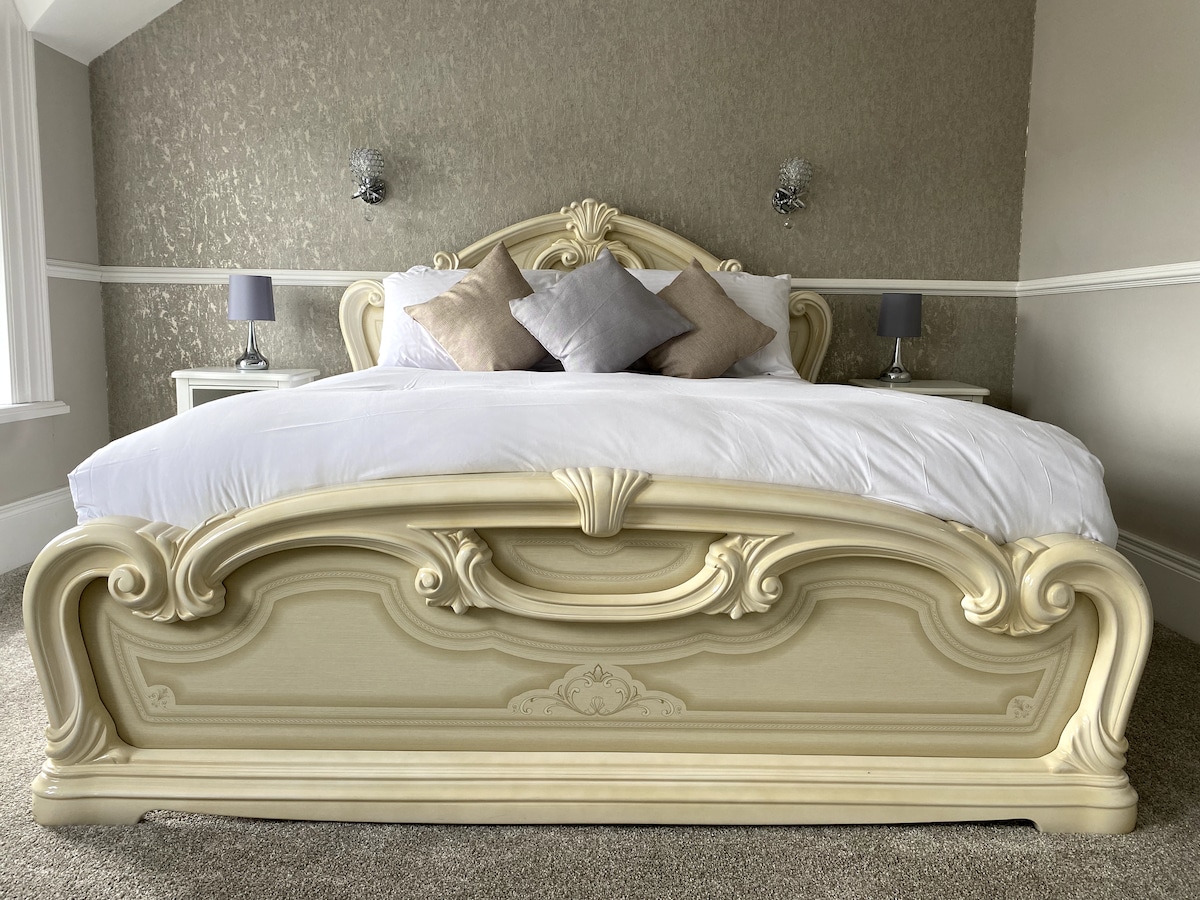 A spacious bed with an ornate headboard is showcased, featuring soft white bedding and decorative pillows. The walls are adorned with a subtle pattern, while bedside tables with lamps are positioned on either side, completing the serene ambiance of the room.