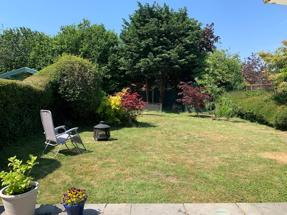 A spacious garden area is visible, featuring manicured grass and various shrubs, including vibrant foliage in shades of green and red. A decorative pot with flowers stands near the patio, while a single lounge chair is positioned on the soft grass beside a fire pit.
