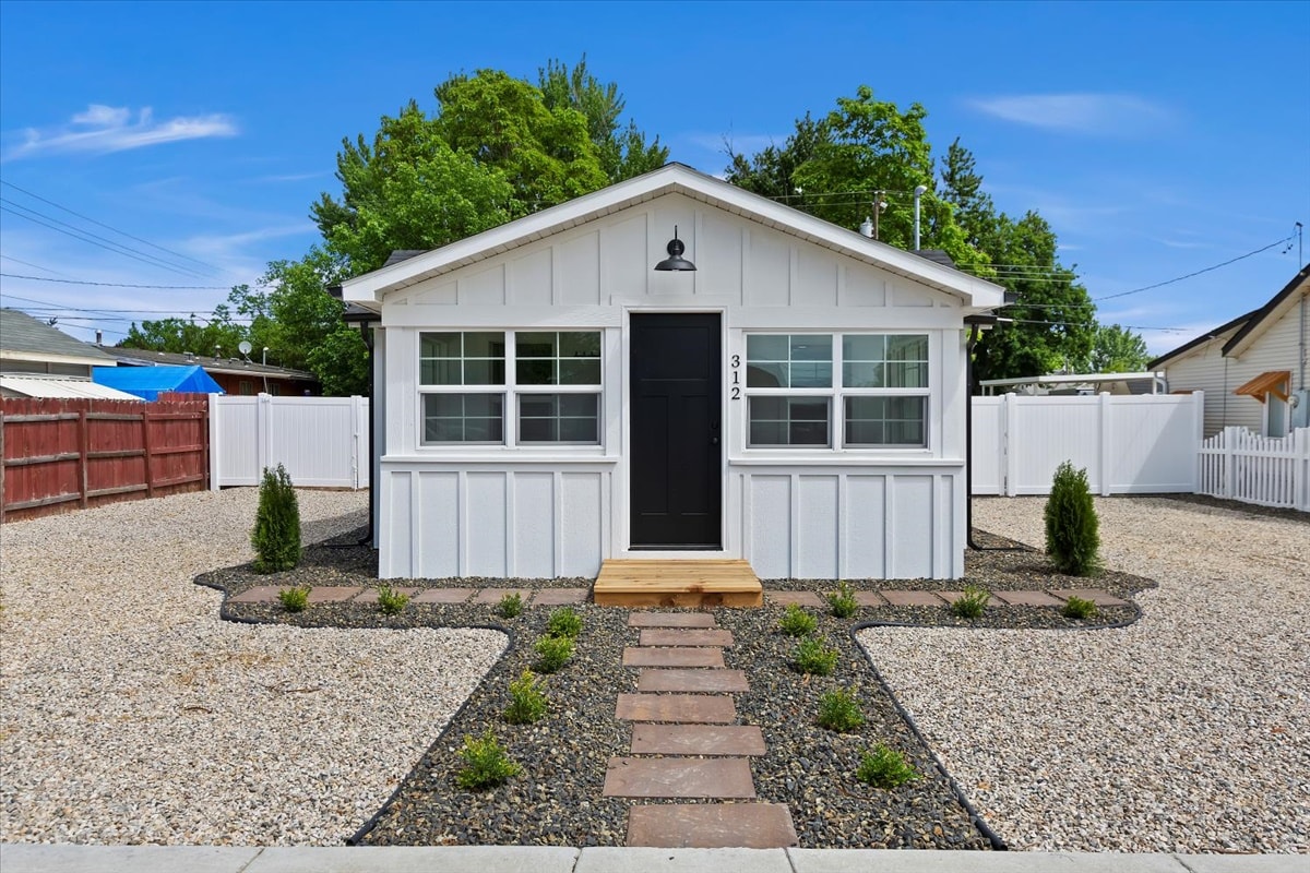 The exterior of the cottage features a clean white facade with a black front door. Surrounding the home, a neatly landscaped area includes a gravel path leading to the entrance, bordered by small plants and shrubs. A large fenced yard creates a private setting.
