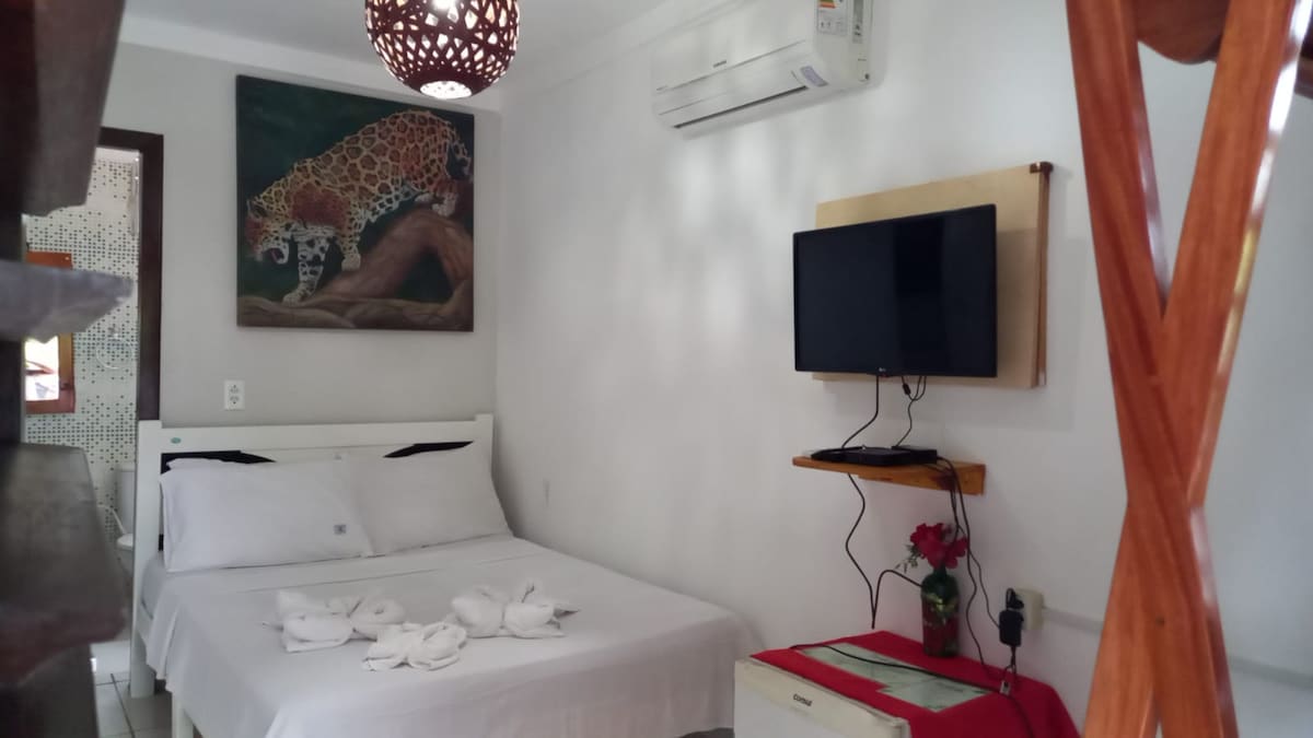 A cozy room is shown with a spacious bed dressed in white linens and decorative towels. A television is mounted on the wall opposite the bed, while a piece of art depicting a leopard adds interest to the space. Air conditioning units and light fixtures provide comfort and ambiance.