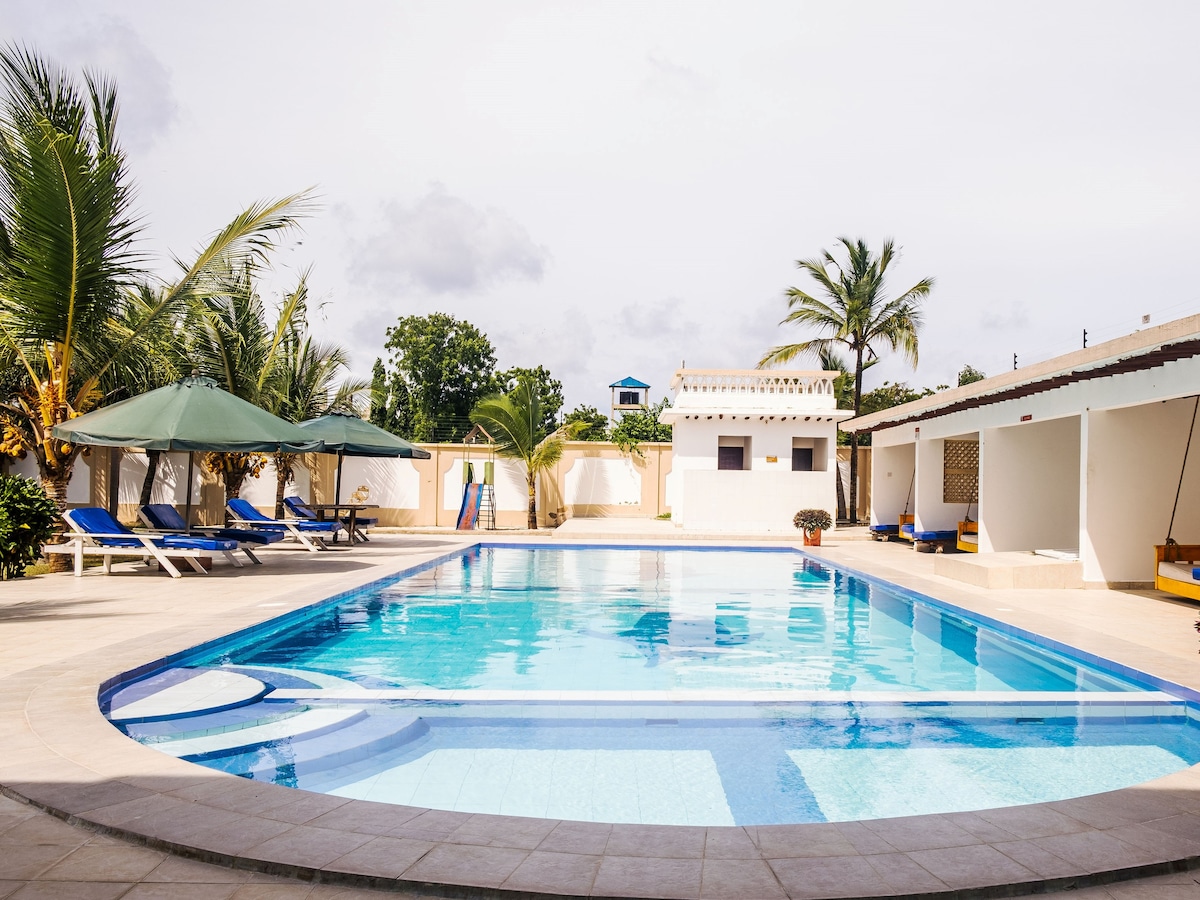The image showcases an outdoor swimming pool surrounded by lounge chairs under large umbrellas. Palm trees provide a tropical vibe, while a small structure is visible in the background. The pool area is well-maintained, promoting a relaxing atmosphere for guests.