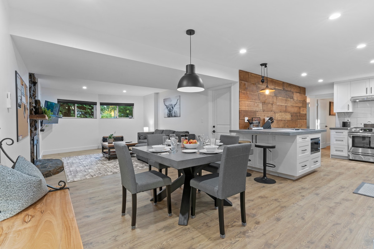 A modern dining area features a dark wood table set for six, surrounded by upholstered gray chairs. An island with bar stools separates the dining space from the kitchen, illuminated by pendant lighting. Natural light enters through large windows, creating a bright and inviting atmosphere.
