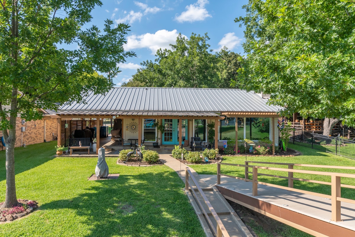A charming cottage exterior is showcased, featuring a welcoming front porch with seating and lush greenery. A paved pathway leads to a wheelchair-accessible ramp, connecting to the dock. The structure is complemented by a metal roof and surrounding well-maintained landscaping.