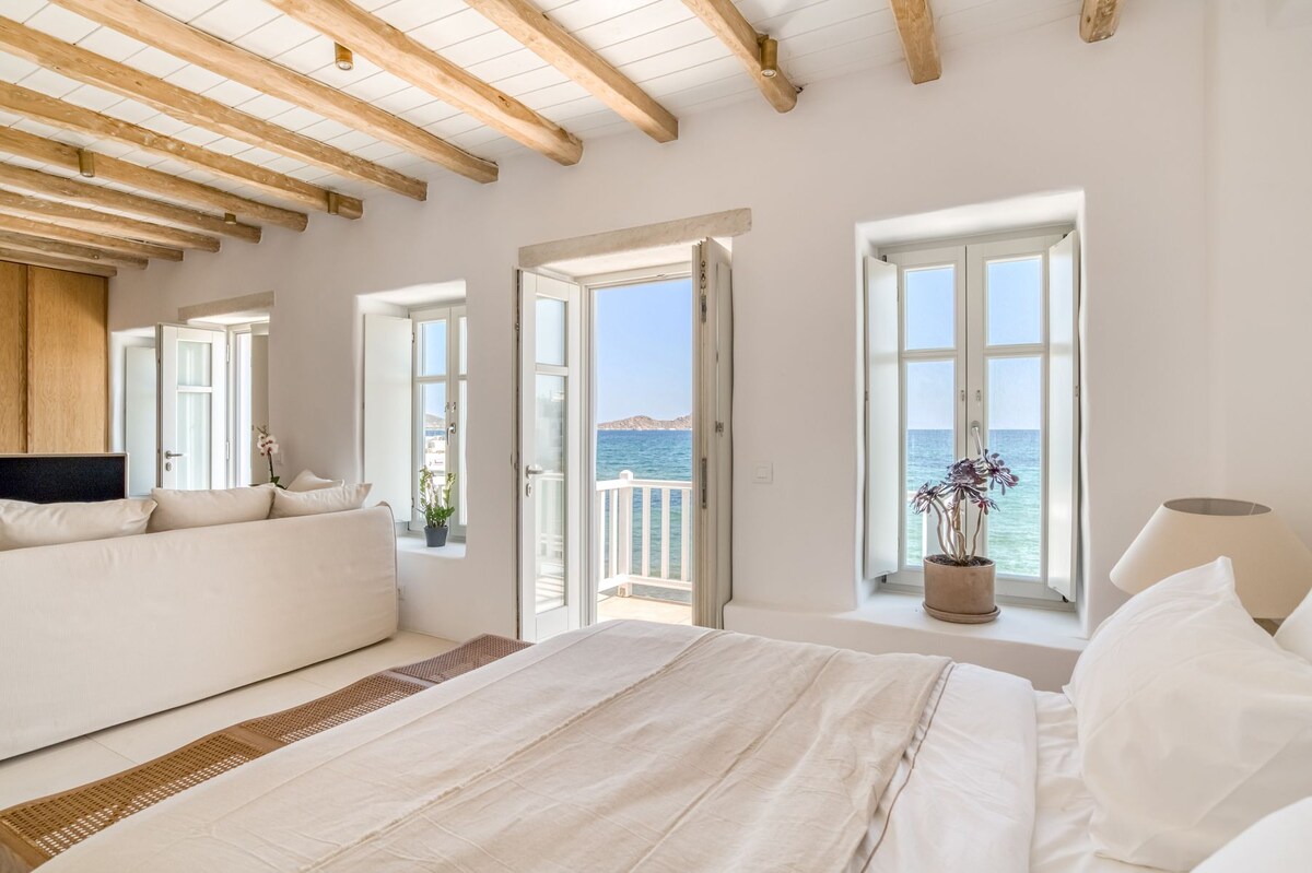 A bright and airy bedroom features a spacious bed with neutral bedding. Natural light floods the room through multiple windows, which offer a view of the sea. Light wooden beams create an open feeling, while a stylish sofa adds comfort to the space.