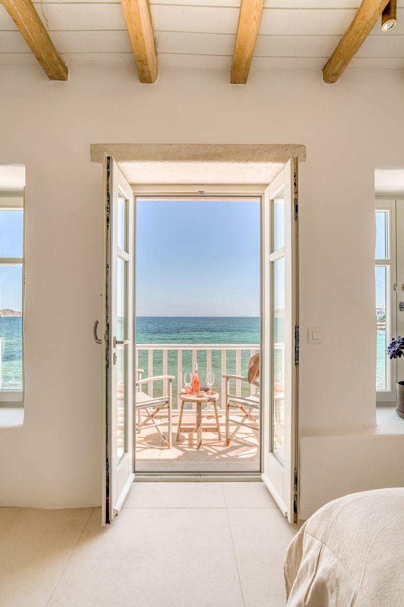 Two white doors stand open, revealing a balcony with two chairs and a small table set for two, overlooking the sparkling blue sea. The light-filled room features large windows on either side, highlighting a serene coastal view.
