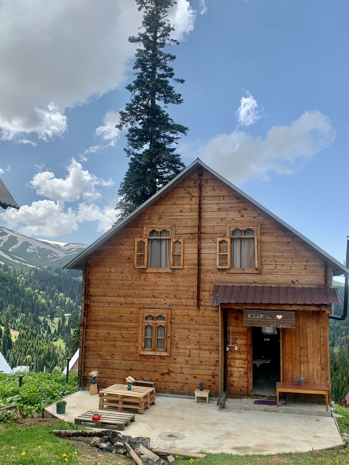The wooden exterior of the unique house is complemented by neatly arranged window shutters and potted flowers by the entrance. Surrounding mountains and trees can be seen in the background, with a partly cloudy sky above.