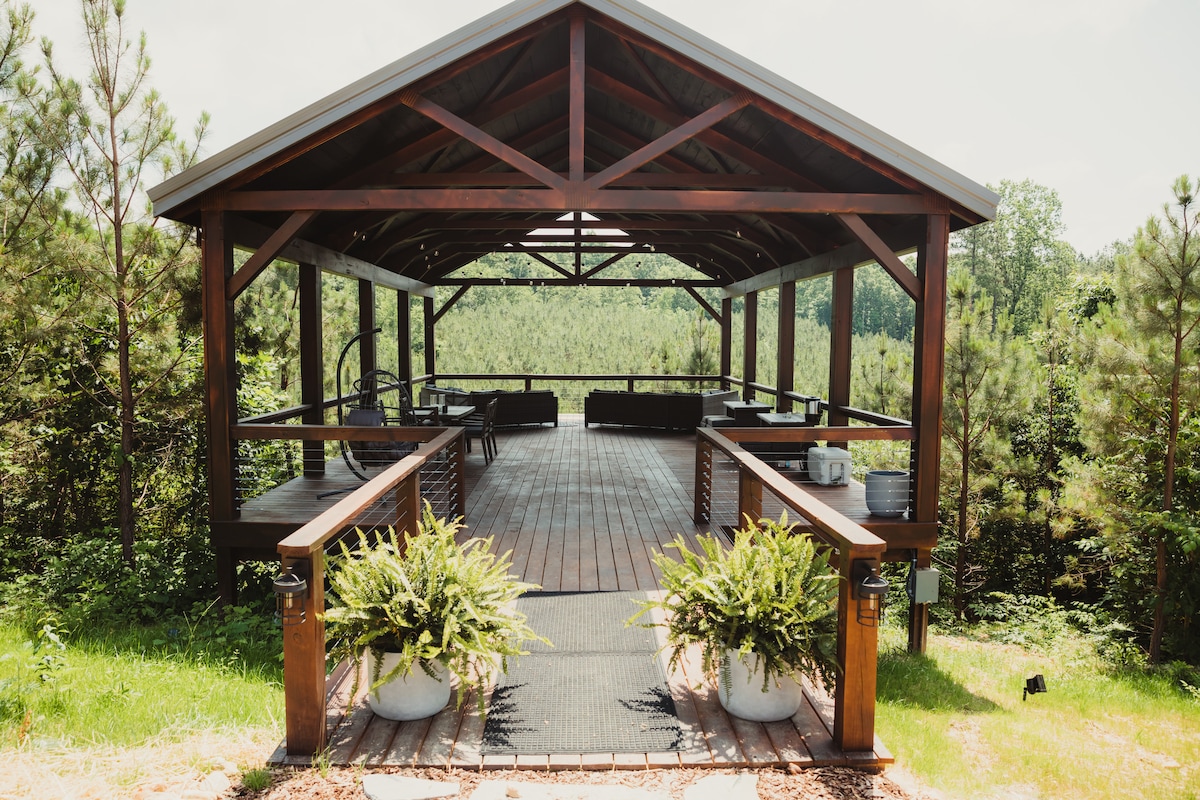 A spacious covered pavilion stands amidst greenery, featuring wooden beams and a large open area. Outdoor sectional sofas and firepit tables surround a central space, while planters with lush foliage add a touch of nature. The structure offers views of the surrounding pines.