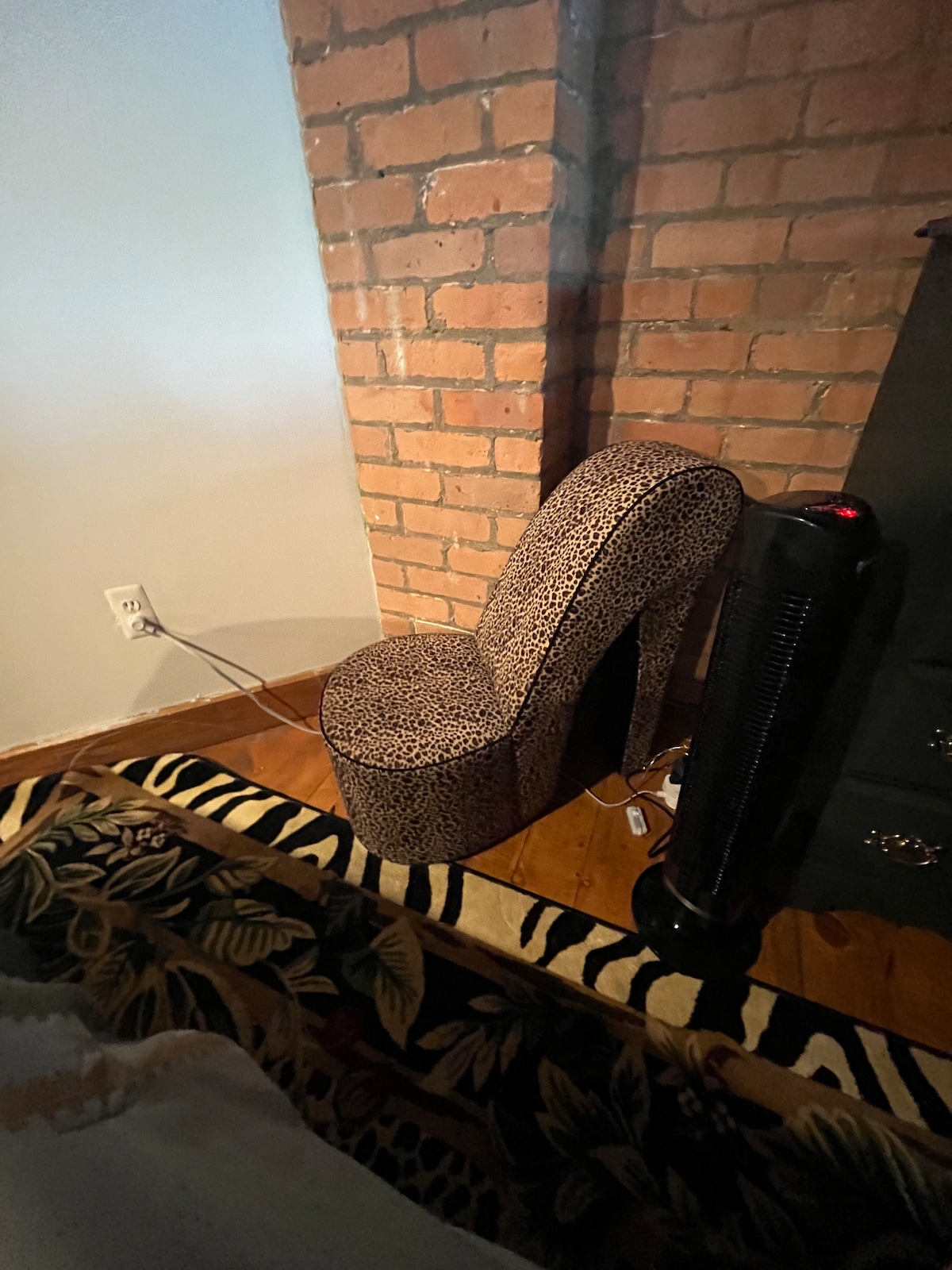 A unique chair with a leopard print design is positioned against a brick wall. The chair’s distinctive shape adds an artistic touch, while a heater and a power cord can be seen nearby. A patterned rug featuring animal prints covers the wooden floor.