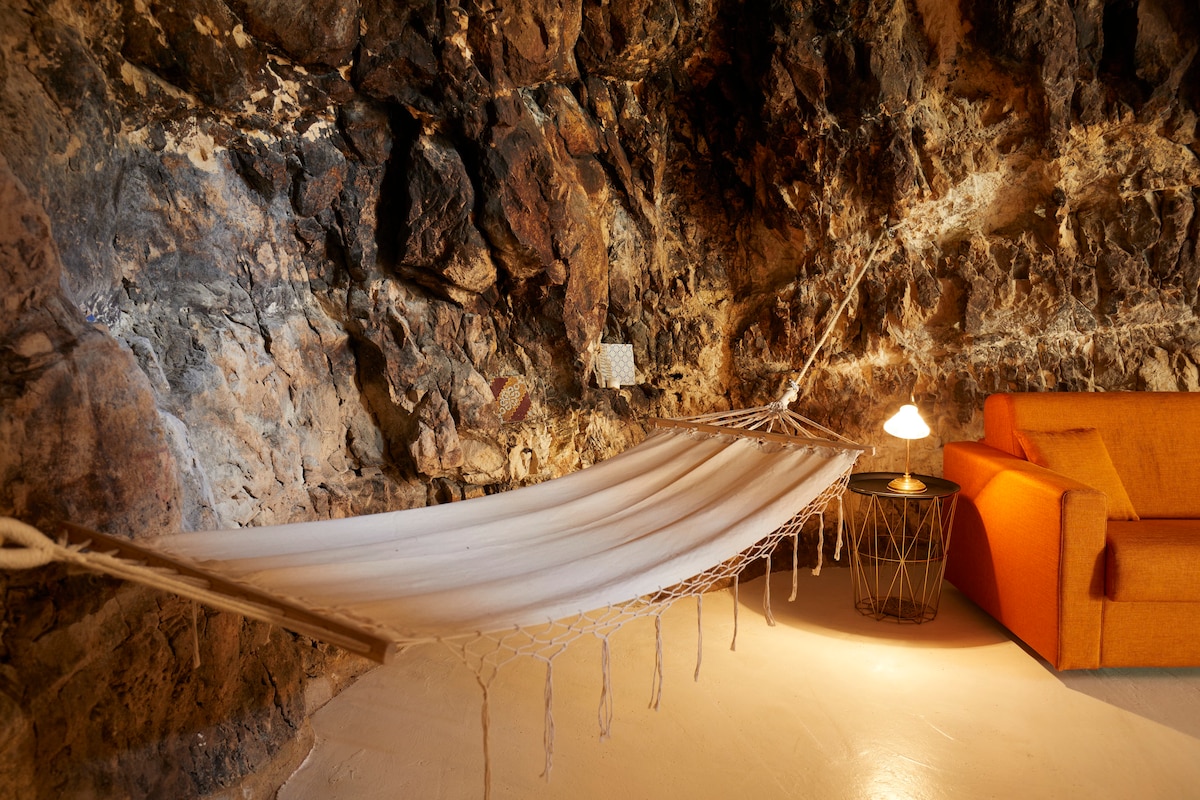 A hammock is suspended between the rugged stone walls of a cave-like interior. An orange couch is positioned nearby, accompanied by a small round table topped with a soft-glow lamp, creating a cozy relaxation space.