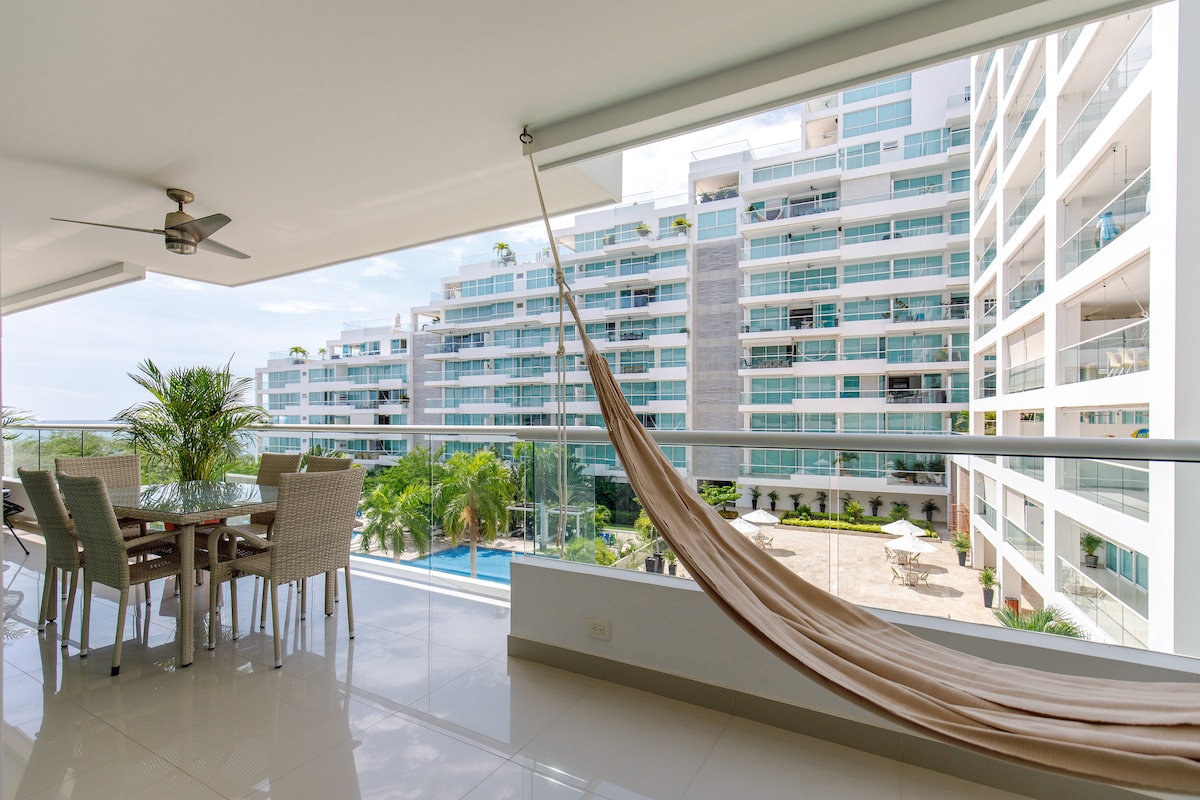 A spacious balcony features a dining area with a glass-top table surrounded by wicker chairs. A light-colored hammock is hung nearby, with views of landscaped grounds and a swimming pool in the distance. Large glass doors allow natural light to illuminate the indoor space.