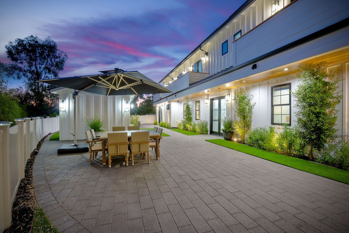An outdoor entertainment space features a large umbrella shading a dining table surrounded by comfortable chairs. The carefully manicured lawn and plants provide a lush backdrop, while the property is illuminated by soft lighting, creating a welcoming atmosphere during twilight.