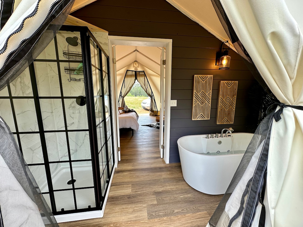 The interior of the safari tent features an open layout with a spacious freestanding tub and a modern walk-in shower. Light-colored walls and flooring enhance the bright atmosphere, while large entryways connect the bathing area to the sleeping space. Natural light filters through the tent openings.