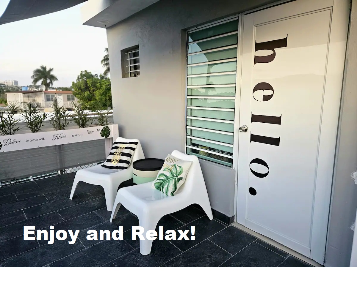 A modern balcony area features two minimalist white chairs, accompanied by a small round table. A door with a friendly greeting is visible, while greenery and palm trees can be seen in the background, enhancing the relaxing outdoor space.