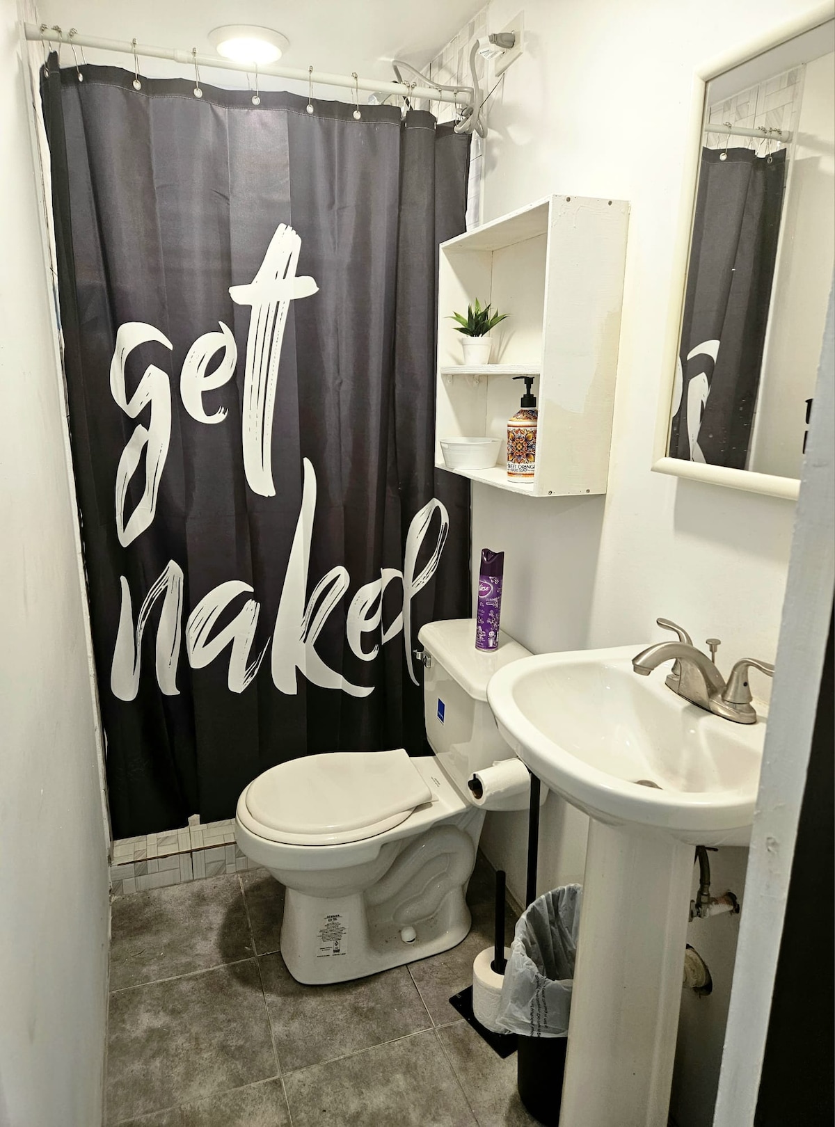 A compact bathroom features a white toilet and a pedestal sink. A decorative shelf holds small plants and toiletries. The shower area is enclosed by a dark curtain with bold lettering, while gray tiles adorn the floor, contributing to a modern aesthetic.