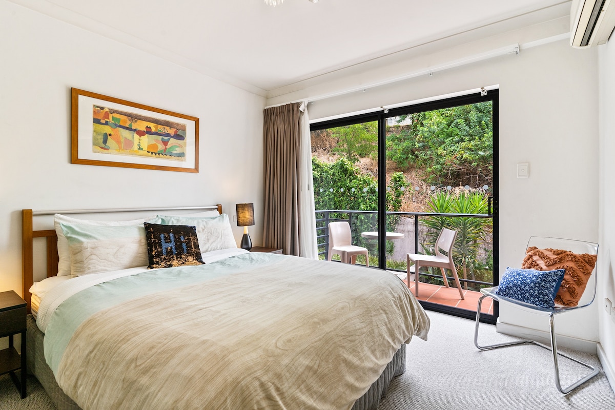 A spacious bedroom features a large bed with soft linens, complemented by a framed artwork above the headboard. Large sliding doors lead to a private balcony with outdoor seating and lush greenery in view, enhancing the connection to nature.