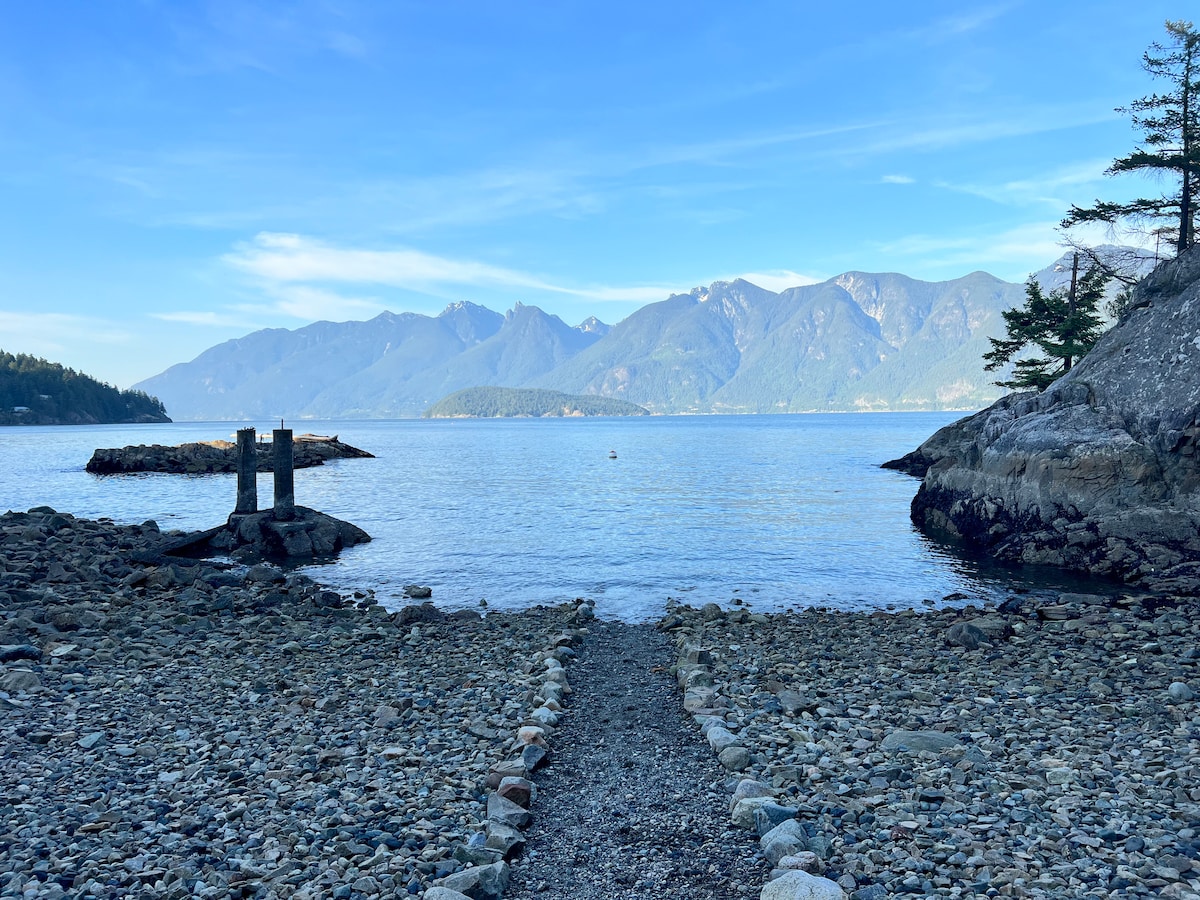 A rocky beach scene features a pebble pathway leading to calm waters. Gentle waves lap against the shore, under a clear blue sky with distant mountains creating a serene backdrop. The unique rock formations on either side emphasize the natural beauty of the coastal landscape.