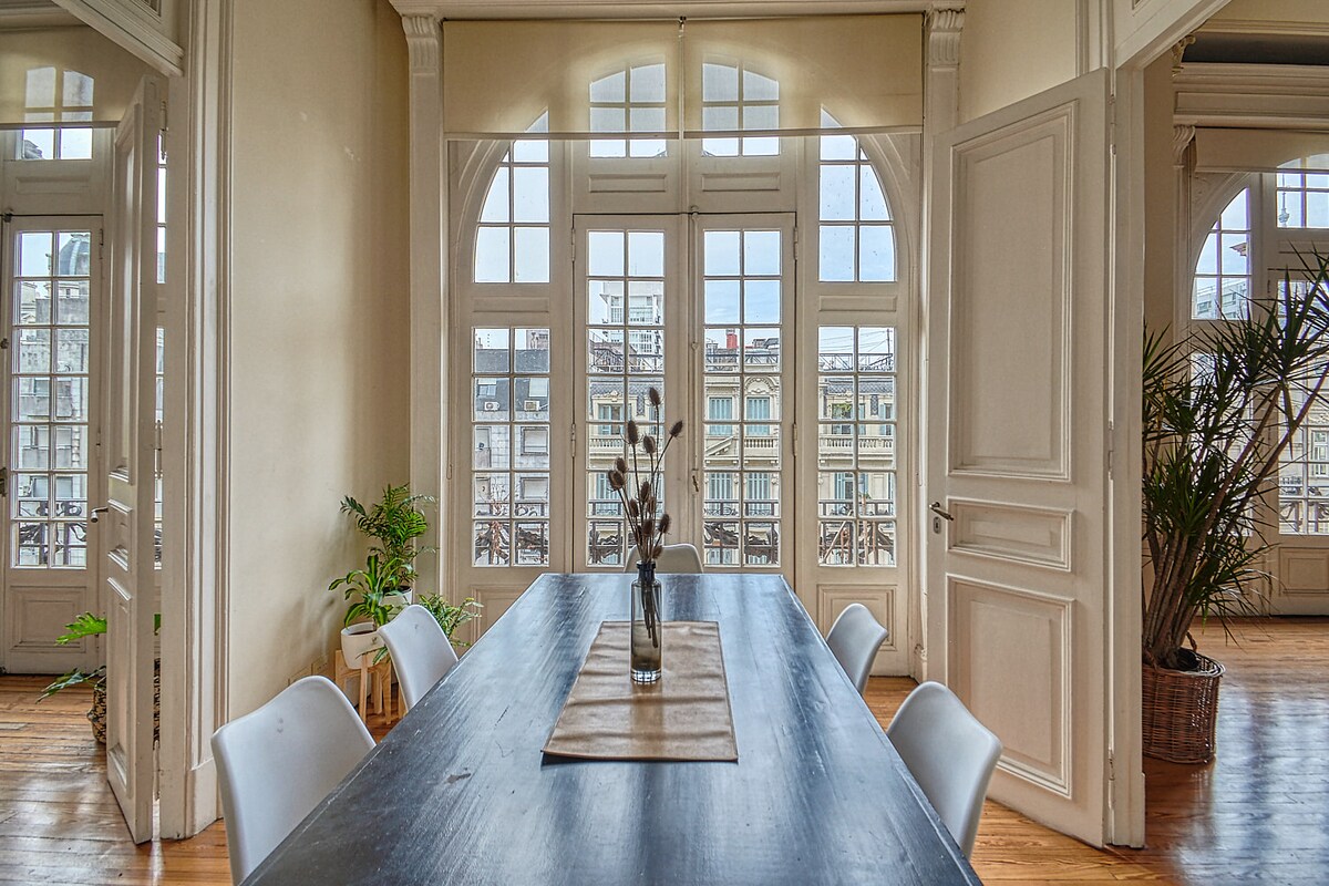 An elegant dining area features a long wooden table surrounded by white chairs. Large, arched windows allow natural light to fill the space, offering views of the surrounding cityscape. Green plants are positioned near the windows, enhancing the room's ambiance.