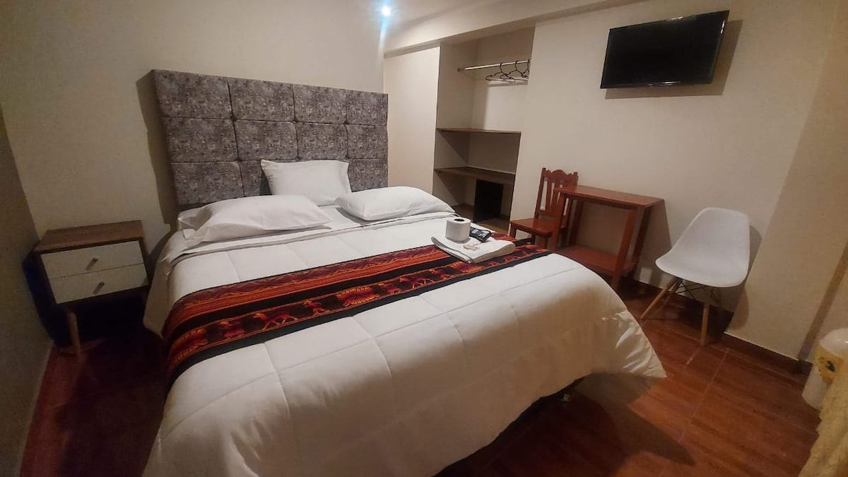 A cozy bedroom features a spacious queen bed dressed in white linens, accented by a decorative woven throw. A small side table and a chair are present, along with a wall-mounted TV and open closet space, providing functionality. Warm lighting enhances the inviting atmosphere.