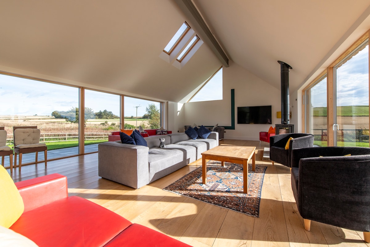 A spacious living area features high ceilings with skylights, allowing natural light to illuminate the warm wood flooring. Comfortable seating is arranged around a coffee table, with a large television mounted on the wall. Expansive windows provide views of the surrounding countryside.