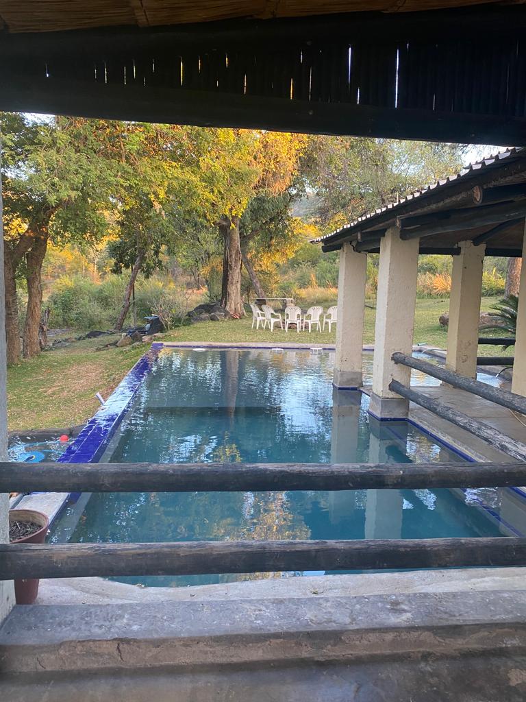 An infinity splash pool is viewed from a covered patio, surrounded by lush trees and scenic greenery. Several outdoor chairs can be seen nearby, inviting relaxation in the serene environment. The tranquil water reflects the natural surroundings, enhancing the calm atmosphere.