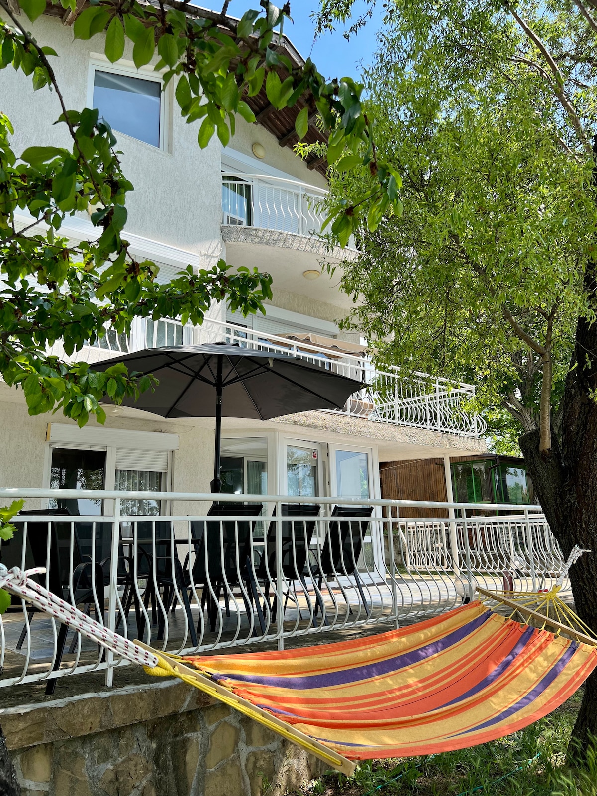 A spacious balcony area is visible, featuring an umbrella over a set of black outdoor furniture. A colorful striped hammock is draped between trees, inviting relaxation. Lush greenery surrounds the space, enhancing the overall calm atmosphere of the outdoor setting.
