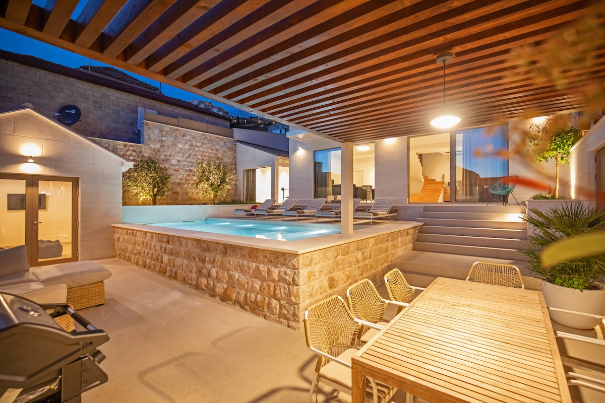 An outdoor area is showcased with a swimming pool surrounded by lounge chairs beneath a wooden pergola. A dining table with wicker chairs is positioned nearby. Soft lighting creates a relaxed ambiance, and the villa's modern architecture blends seamlessly with natural elements.