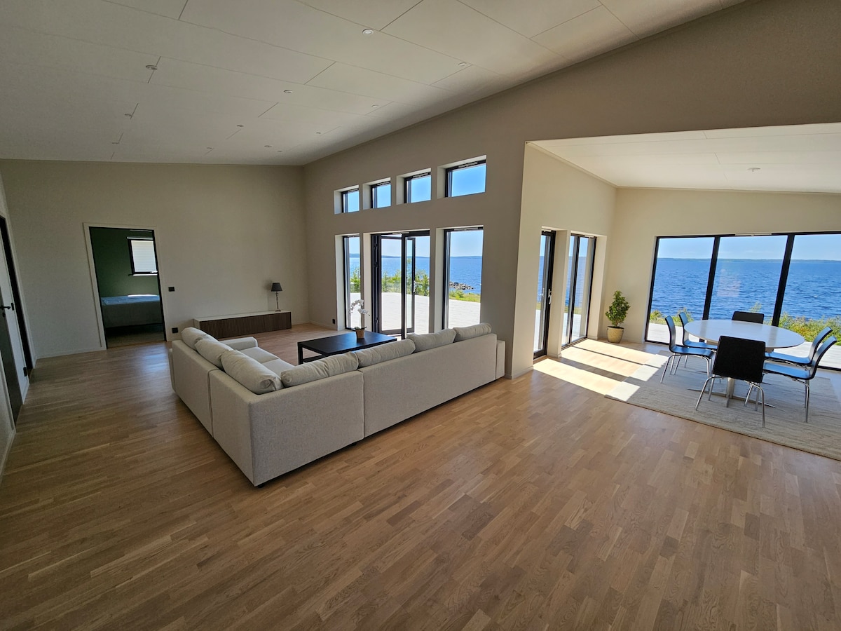 A spacious living area features large windows that provide views of the nearby water. A light-colored sectional sofa is arranged around a dark coffee table. A dining area with a glass table and metal chairs is positioned adjacent to the windows, inviting natural light into the space.