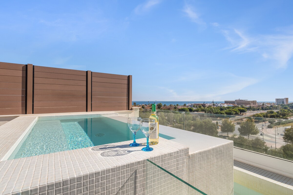 A private plunge pool is situated on a solarium, surrounded by a tiled deck. Two glasses and a bottle of wine are placed on a ledge beside the pool. A clear view of the surrounding landscape, including sea and greenery, is visible in the background.