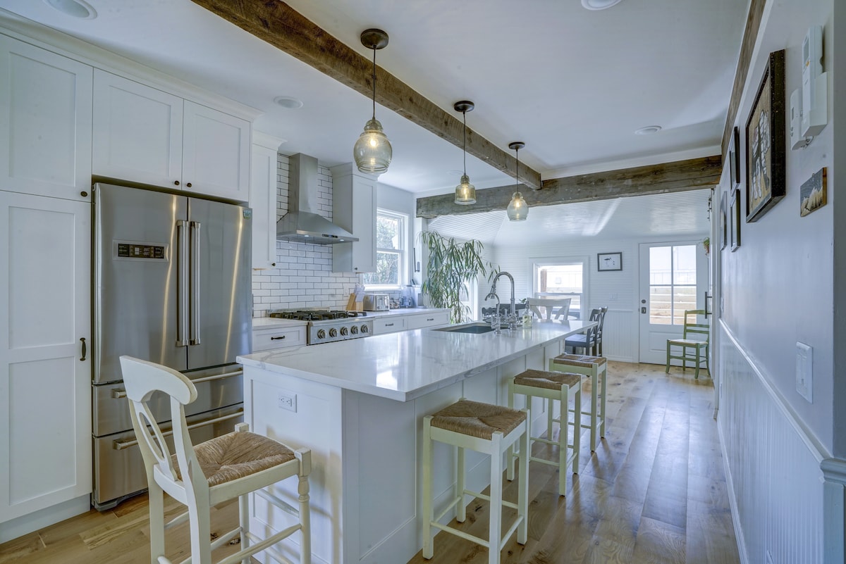 A modern kitchen features white cabinetry and a large central island with bar seating. Pendant lights hang above, illuminating the space. Stainless steel appliances and a tiled backsplash complement the clean design, while a dining area is visible at the end of the room next to large windows.
