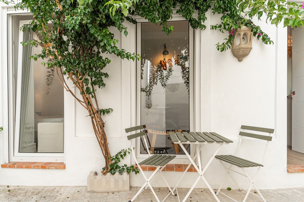 An outdoor seating area features a small, foldable table accompanied by two chairs. Lush greenery climbs along the wall, providing a natural element to the space. Two windows, flanked by light curtains, are visible in the background, contributing to a refreshing ambiance.