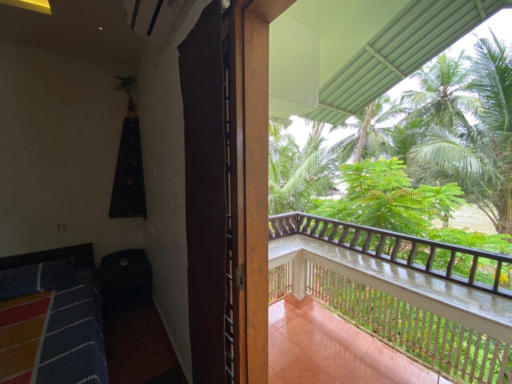 A sliding door opens to a private balcony offering views of lush greenery and palm trees. The interior features minimalistic decor, with a spacious bed visible and natural light filtering in through the open doorway.