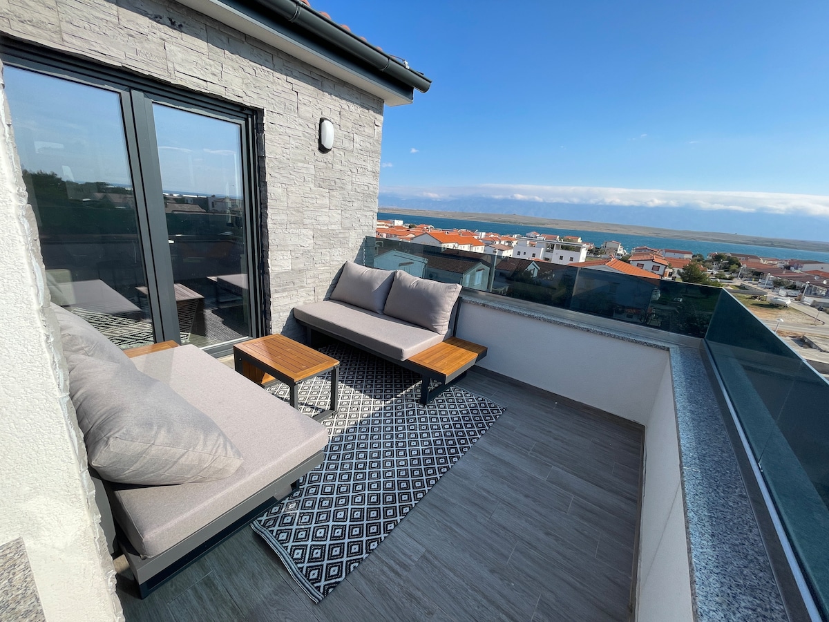 A roof terrace is shown with modern seating arrangements, including two plush lounge chairs and a low wooden table. A patterned rug lies underneath, while a clear view of the sea and surrounding buildings is visible through glass railings, providing an open and airy atmosphere.