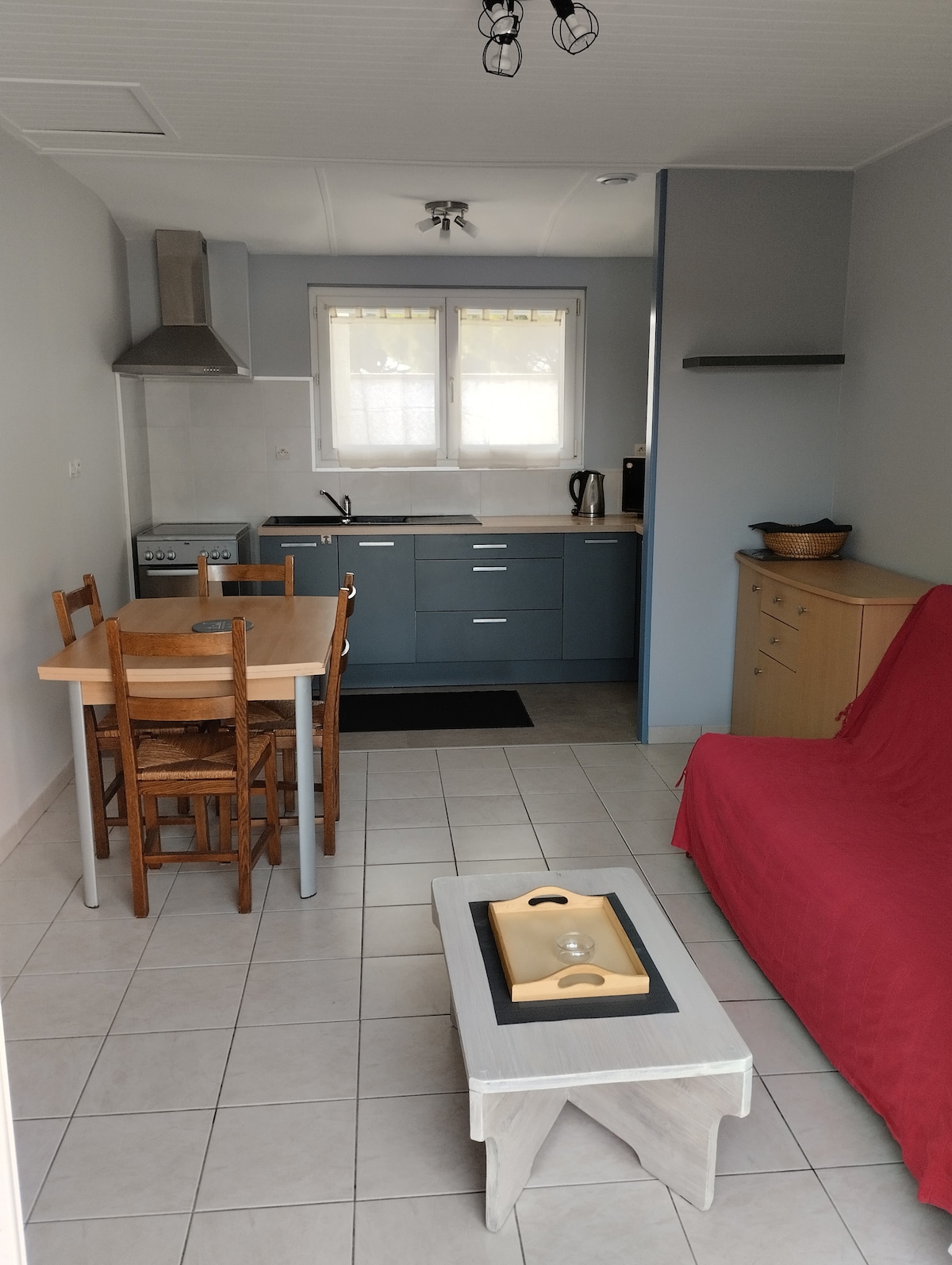 A compact kitchen area is equipped with a stove and sink, accompanied by light-colored cabinetry. A wooden dining table with four chairs sits beside a red couch. Natural light enters through nearby windows, casting a soft glow on the tiled floor.