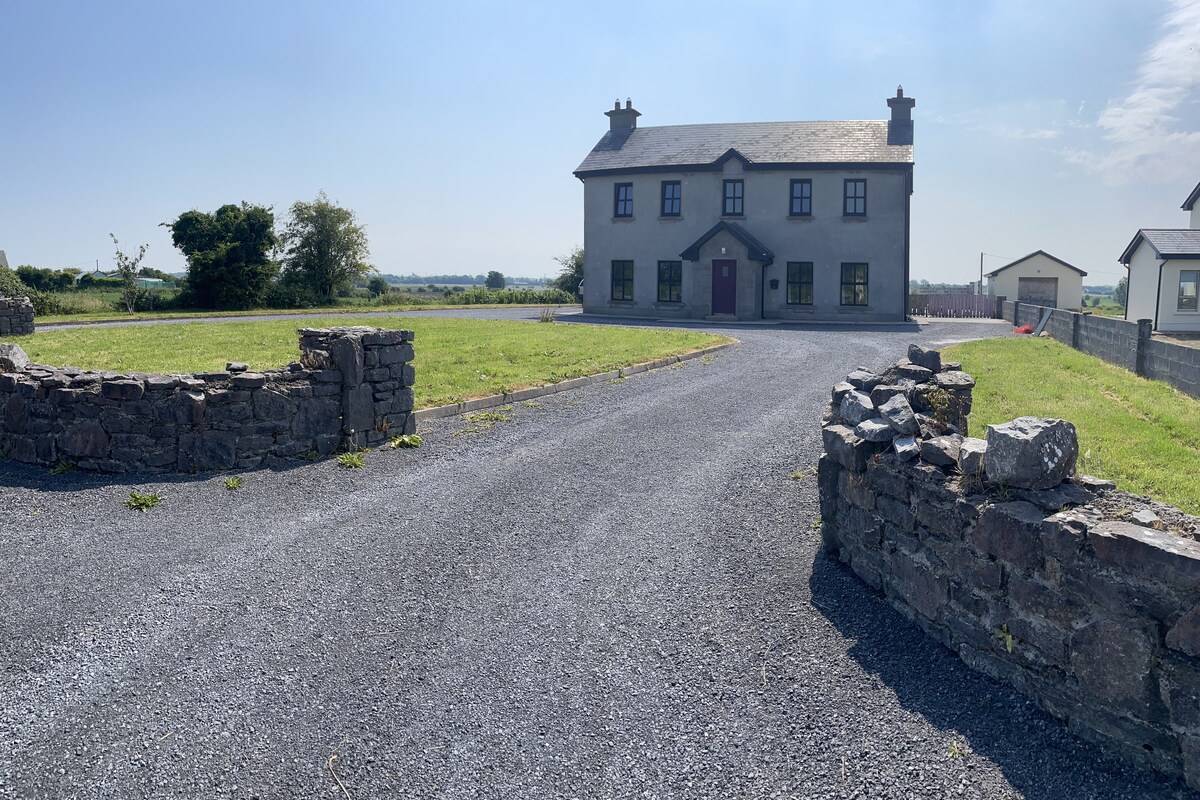 A large two-story house is set back from a gravel driveway, surrounded by a green lawn and open fields. Stone walls frame the entrance, complemented by clear blue skies overhead. The exterior features simple architectural lines and a muted color palette, enhancing its rustic charm.