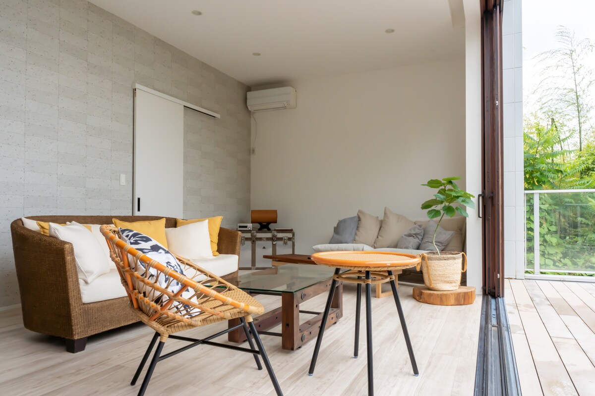A sunlit living area features a comfortable sofa adorned with yellow and grey cushions. A glass coffee table is center stage, complemented by a round wooden table and a rattan chair. Large glass doors provide access to a lush outdoor space, enhancing the openness of the room.
