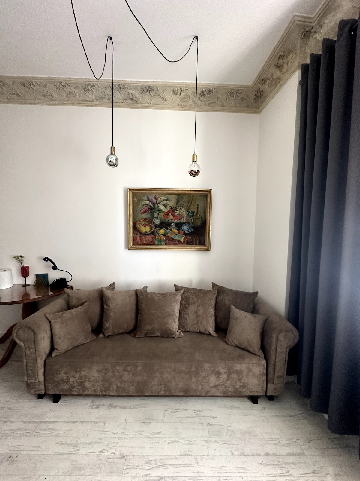 A comfortable seating area is presented with a gray upholstered sofa adorned with multiple cushions. A colorful piece of artwork hangs on the wall above the sofa, while a small table with a lamp is positioned nearby. Floor-to-ceiling curtains frame the window, contributing to the room's ambiance.