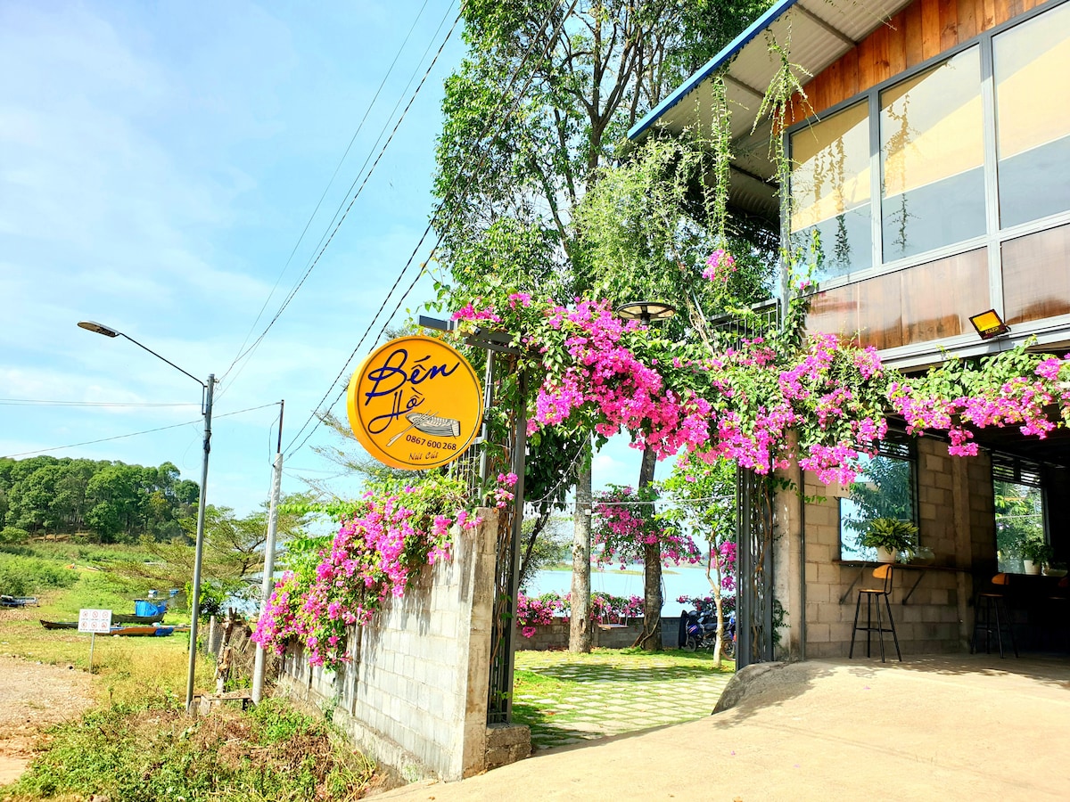 The entrance features a vibrant sign with the name of the property, surrounded by lush bougainvillea flowers. The building showcases large glass windows and is framed by greenery, with an inviting pathway leading towards a serene water view.