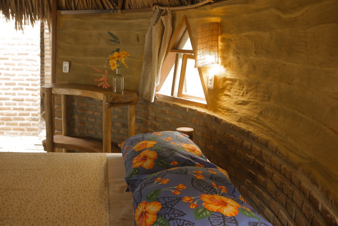 An interior view reveals a unique organic design featuring curved earthy walls and a straw thatched ceiling. A bed adorned with a floral patterned blanket rests against the wall, complemented by a wooden side table. Natural light filters through an angled window, creating a warm glow.
