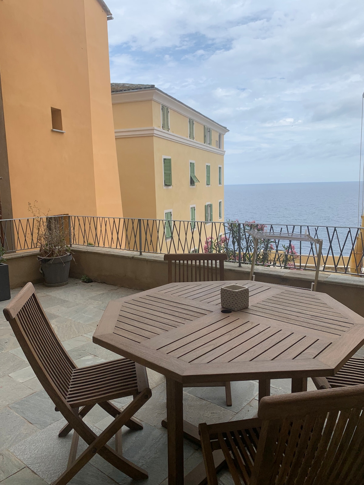 An outdoor terrace features a wooden octagonal table surrounded by four slatted chairs. Potted plants enhance the space, and a view of the sea is visible beyond the railing. The surrounding buildings display pastel-colored facades under a cloudy sky.