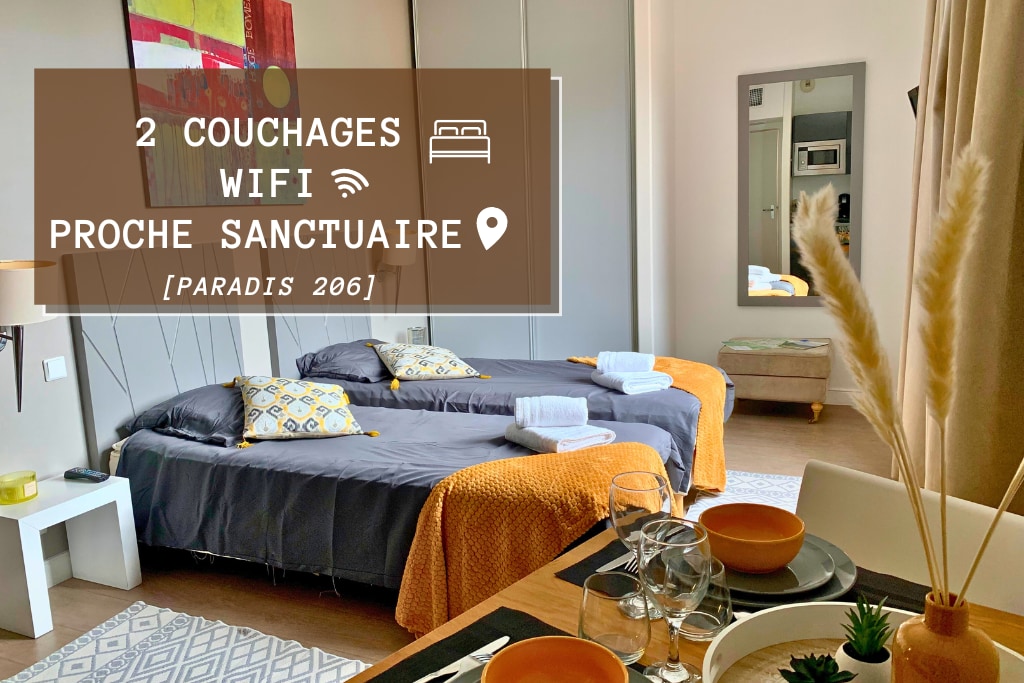 The studio features two single beds dressed in dark linens, complemented by decorative cushions. A dining area with neatly arranged tableware is included, and light streams in through large windows, enhancing the bright ambiance of the space.