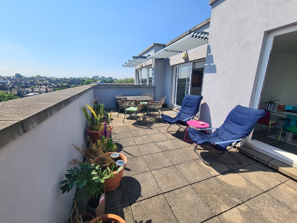 A private terrace is showcased, featuring two blue lounge chairs and a small round table. Various potted plants add greenery to the space. The setting allows for outdoor relaxation with views of the surrounding area under clear blue skies.