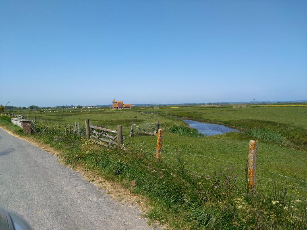 A scenic view captures expansive green fields under a clear blue sky. A small stream meanders through the landscape, and a rustic wooden fence lines the roadside. A charming building can be seen in the distance, complementing the natural surroundings.