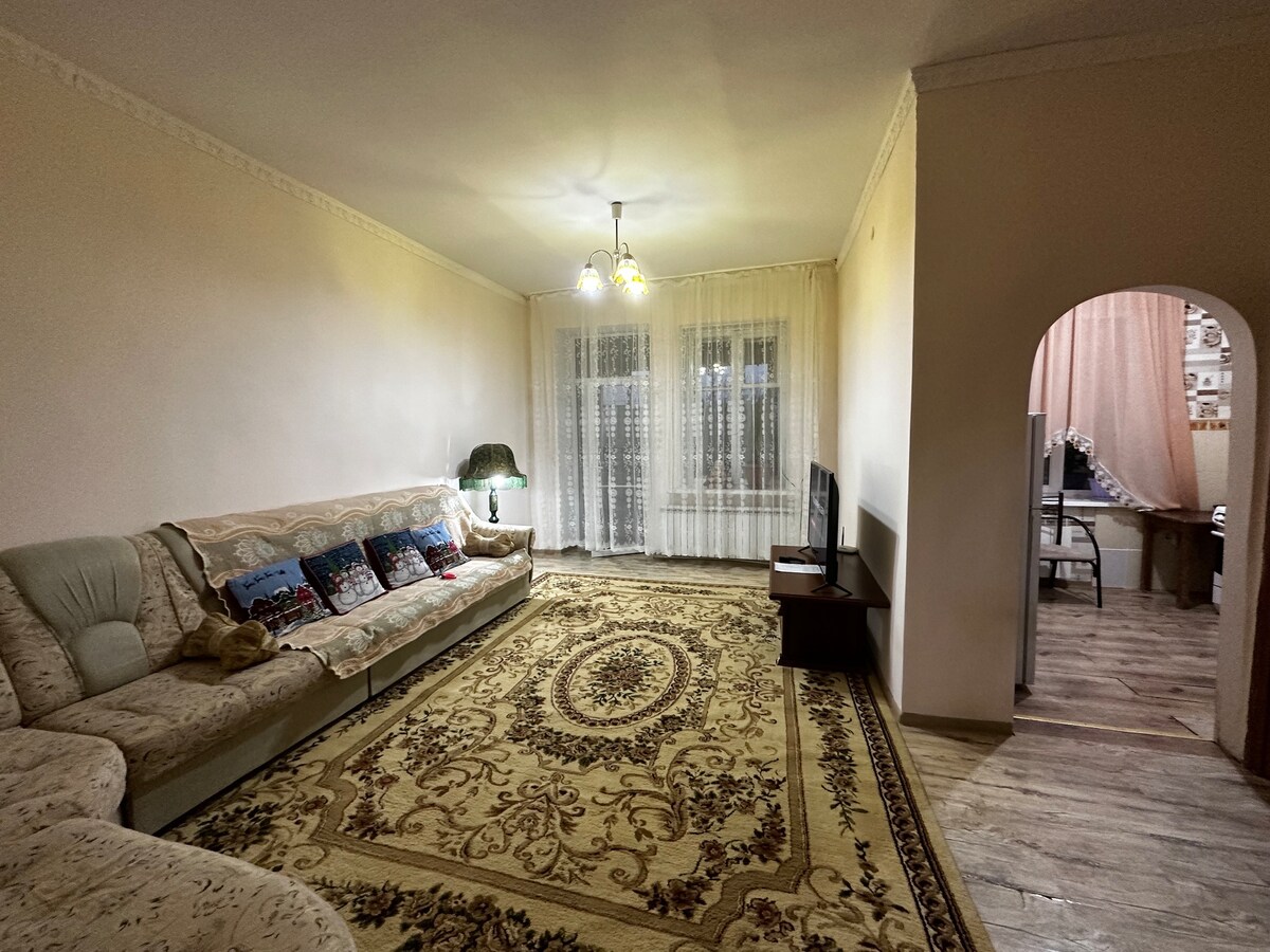 A spacious living area features a plush sofa adorned with decorative pillows, positioned on a patterned area rug. Natural light filters through sheer curtains covering the large windows. A flat-screen television is placed on a wooden stand, with an archway leading to another room in the background.