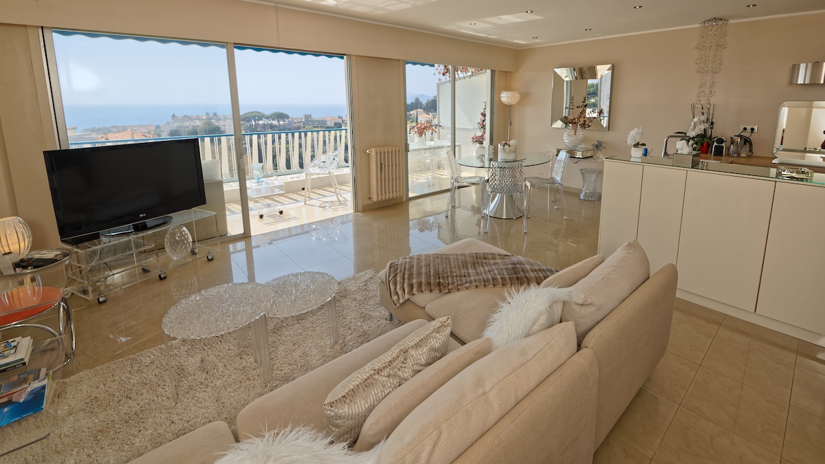 A bright living area is showcased with large glass windows offering a view of the sea. The space features a comfortable beige sofa, a modern television, and a dining area with clear chairs. Natural light fills the room, enhancing the tranquil ambiance.
