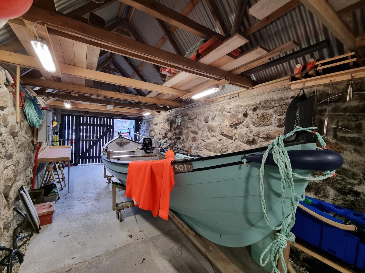 An old boathouse interior is visible, showcasing wooden beams and stone walls. Two boats are prominently displayed, with one featuring a vibrant orange cover. Tools and equipment are neatly arranged along the sides, while a workbench stands ready for use near the back.