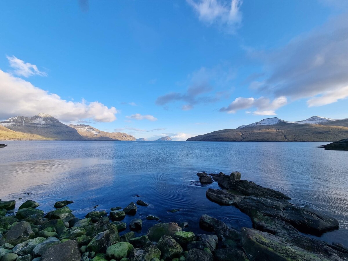 A serene coastal view presents calm waters reflecting the clear blue sky. Rugged mountains rise in the distance, with patches of snow accentuating their peaks. Smooth stones and scattered rocks border the shoreline, inviting exploration of the natural surroundings.