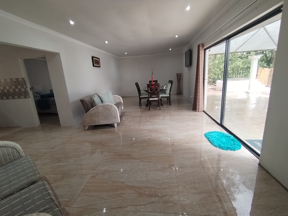 A spacious living area showcases a dining table surrounded by four chairs. Two comfortable sofas are positioned nearby on polished tile flooring. Large sliding doors allow natural light to enter while providing views of the outdoor space.